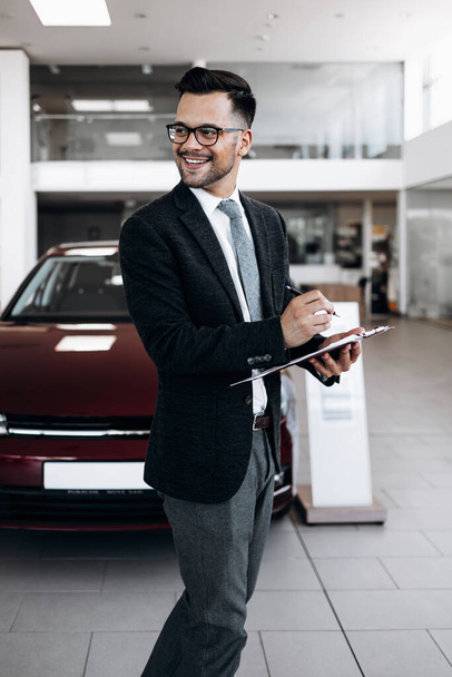 Good looking, cheerful and friendly salesman poses in a car salon or showroom. - Photo, image