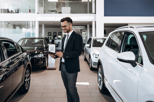 Good looking, cheerful and friendly salesman poses in a car salon or showroom. - Zdjęcie, obraz
