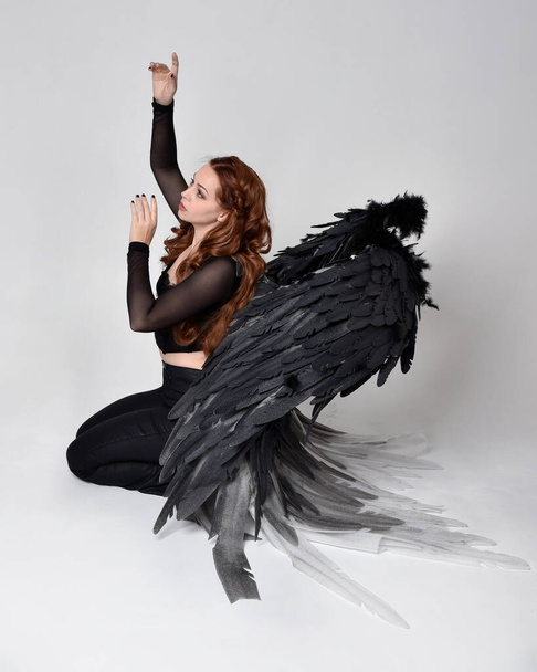 Full length portrait of beautiful woman with long red hair wearing sheer corset top, leather pants & large black angel feather wings.  Sitting pose with gestural hands reaching out, kneeling on floor. Isolated on white studio background - Photo, image