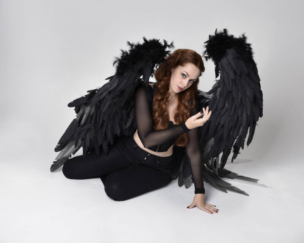 Full length portrait of beautiful woman with long red hair wearing sheer corset top, leather pants & large black angel feather wings.  Sitting pose with gestural hands reaching out, kneeling on floor. Isolated on white studio background - Photo, image