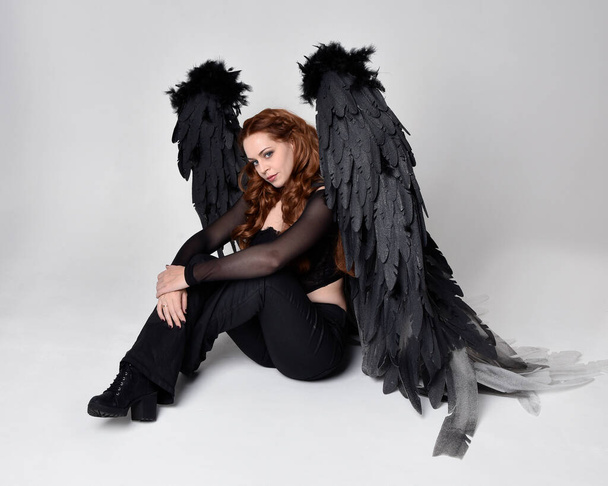 Full length portrait of beautiful woman with long red hair wearing sheer corset top, leather pants & large black angel feather wings.  Sitting pose with gestural hands reaching out, kneeling on floor. Isolated on white studio background - Valokuva, kuva