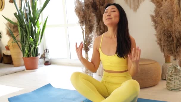 Entspannte junge Asiatin in gelber Sportbekleidung meditiert in Lotusposition und sitzt mit geschlossenen Augen zu Hause auf einer Yogamatte und genießt die Meditation mit den Händen im Gesicht. - Filmmaterial, Video