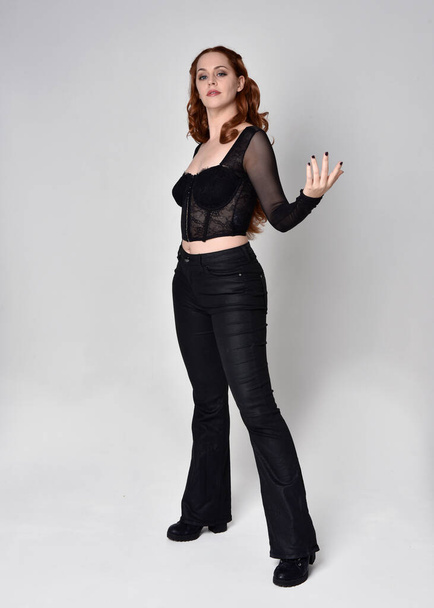 Full length portrait of beautiful woman with long red hair wearing black corset top and leather pants. Standing pose, walking forwards with gestural hands reaching out. Isolated on white studio background. - Photo, Image