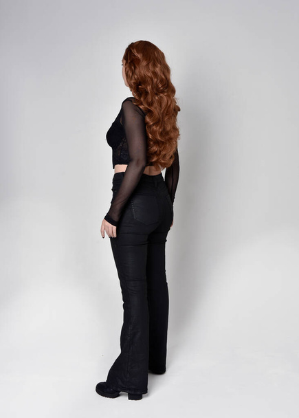 Full length πορτρέτο της όμορφης γυναίκας με μακριά κόκκινα μαλλιά φορώντας μαύρο κορσέ κορυφή και δερμάτινο παντελόνι. Στέκεται όρθιος, κοιτάζει προς τα πίσω, απομακρύνεται από την κάμερα. Απομονωμένο σε λευκό φόντο στούντιο. - Φωτογραφία, εικόνα