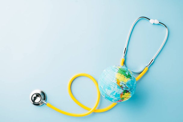 World Health Day Concept. Top view yellow doctor stethoscope and world globe isolated on pastel blue background with copy space, Save world day, Environment Green Earth, Health care and medical - 写真・画像