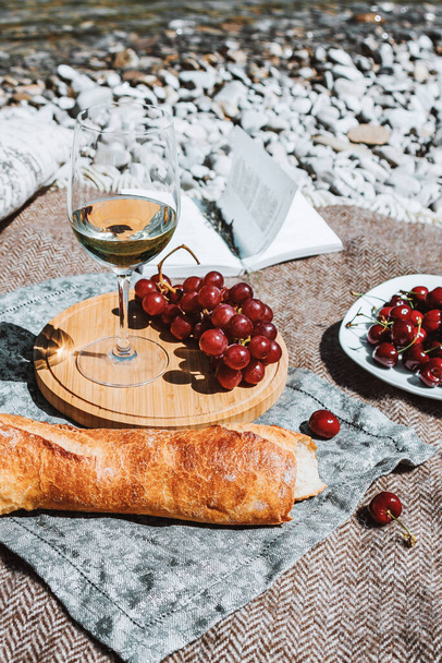 Summer romantic rustic picnic on the coast with white wine glass, baguette, cherries, grape, open book, pillow. Sea beach. Vacation, rest in Europe. Alcohol drink. Inspiration. High quality photo - Foto, Imagen