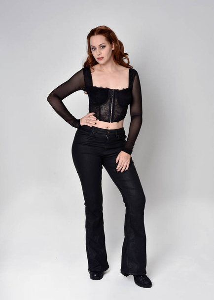 Full length portrait of beautiful woman with long red hair wearing black corset top and leather pants. Standing pose, walking forwards with gestural hands reaching out. Isolated on white studio background. - Foto, imagen