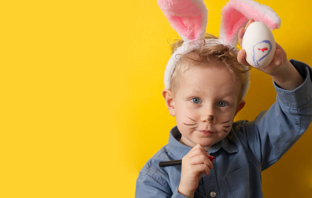 ウサギの耳を持つかわいい男の子は黄色の背景コピースペースでイースターエッグを示しています。高品質の写真 - 写真・画像