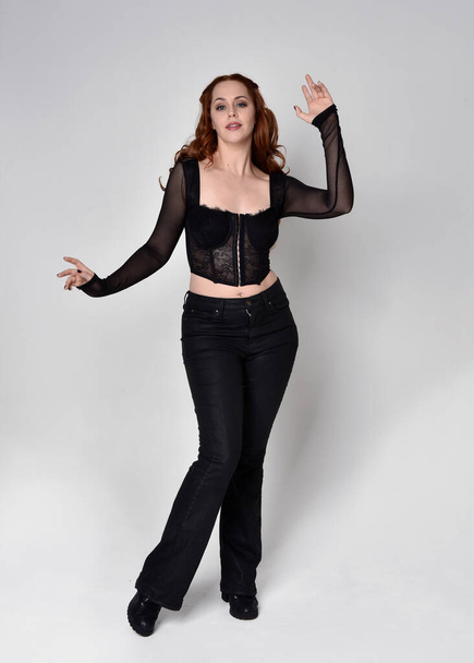 Full length portrait of beautiful woman with long red hair wearing black corset top and leather pants. Standing pose, walking forwards with gestural hands reaching out. Isolated on white studio background. - Foto, Imagen