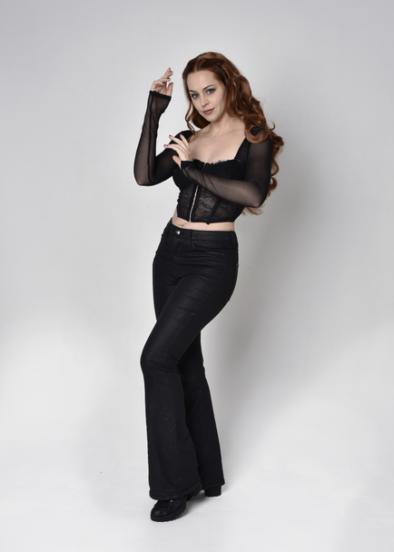 Full length portrait of beautiful woman with long red hair wearing black corset top and leather pants. Standing pose, walking forwards with gestural hands reaching out. Isolated on white studio background. - Fotó, kép