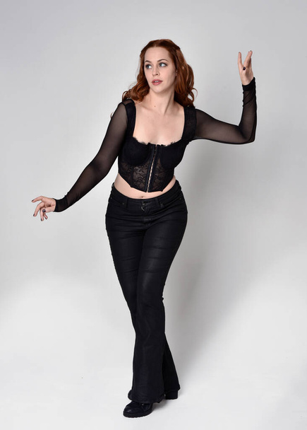 Full length portrait of beautiful woman with long red hair wearing black corset top and leather pants. Standing pose, walking forwards with gestural hands reaching out. Isolated on white studio background. - Φωτογραφία, εικόνα
