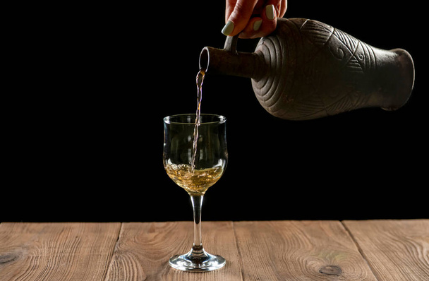 girl hand pouring wine into glass from clay bottle on black background - Photo, Image
