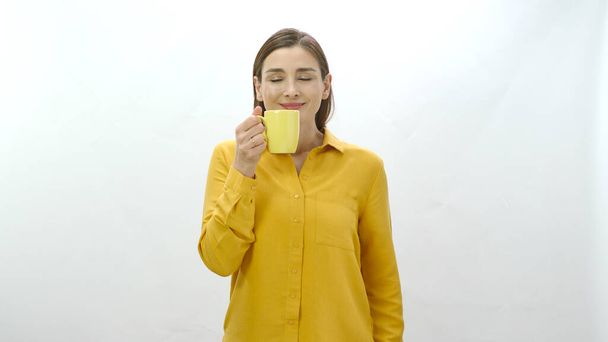 Персонаж портрета молодой женщины, пьющей чашку кофе, чёрный или зелёный чай. Молодая здоровая женщина нюхает чашку кофе или чая на белом фоне, указывая на камеру - Фото, изображение