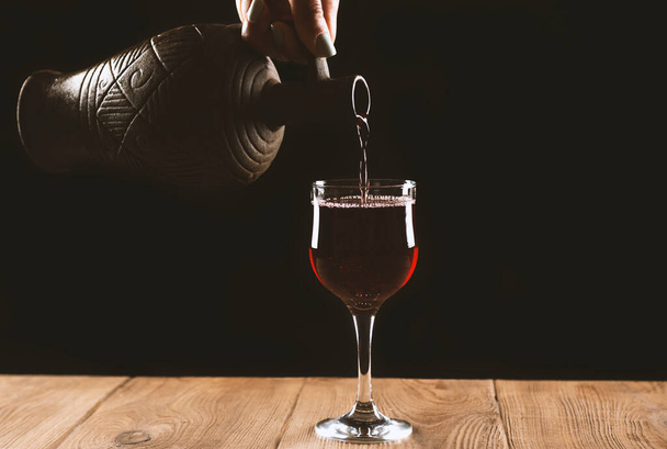 girl hand pouring red wine into glass from clay bottle on black background - Photo, image