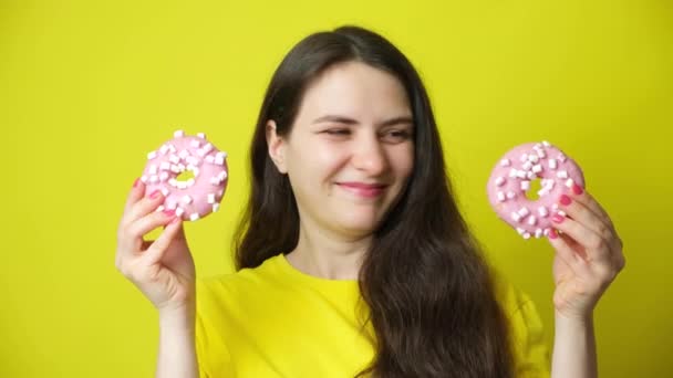Eine lustige brünette Frau spielt mit Donuts, setzt sie wie eine Brille auf Augenhöhe und zeigt ihre Zunge - Filmmaterial, Video