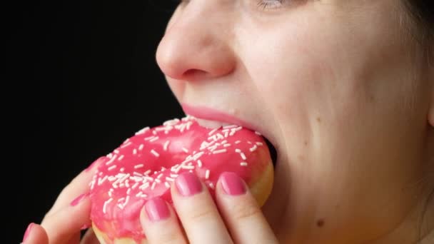 Een vrouw eet een heerlijke rode donut, bijt en kauwt er smakelijk op, close-up. Zoet voedsel met veel calorieën, diabetes. - Video