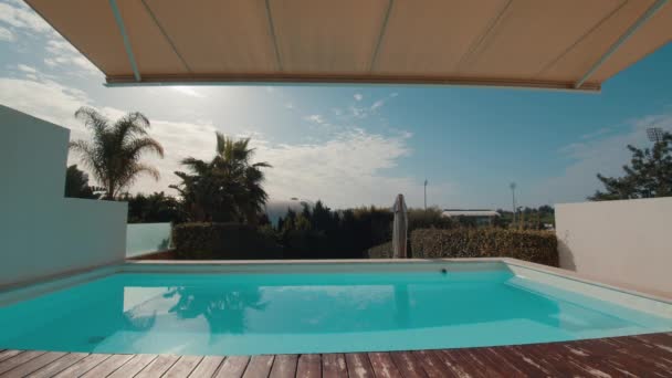 View of a pool surrounded by lush vegetation and palm trees at a villa in Portugal. A tent cover is slowly opened, allowing the bright sun to shine in - Video