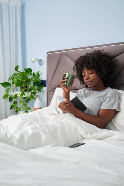 African young woman reading a book and drinking coffee in bed in the morning. - Foto, imagen