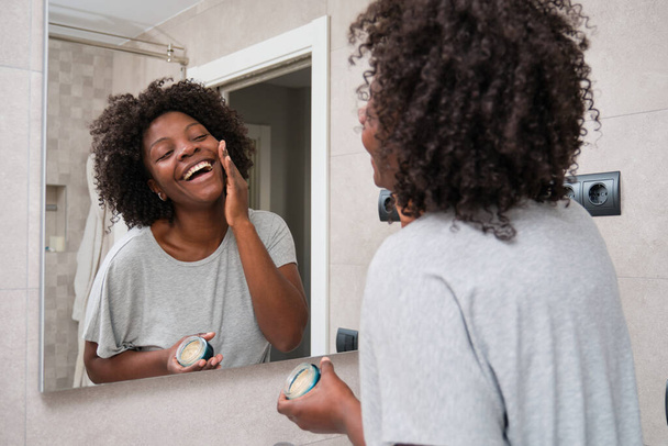 Smiling african young woman applying eco-friendly facial cream in bathroom. - Foto, immagini