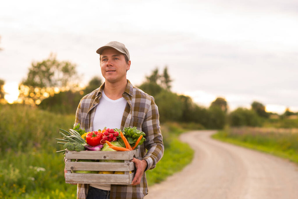 Farmer with a vegetable box in front of a sunset agricultural landscape. Man in a countryside field. The concept of country life, food production, farming and country lifestyle. - Valokuva, kuva