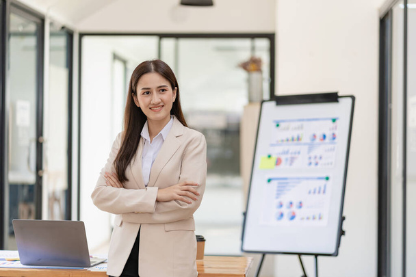 Young asian female accountant standing with arms crossed in front of desk with laptop and financial documents in background in office - Фото, зображення