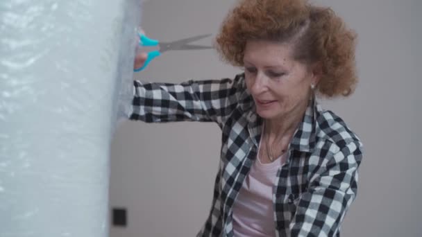 Aged woman bought new mattress and is unpacking it from factory package in new empty apartment. Moving and furniture fitting in new home. Elderly lady opens mattress during move to newly property.  - Footage, Video