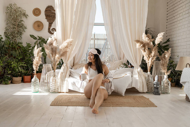 Imagem da bela jovem mulher asiática em lingerie e em uma máscara de dormir sorrindo e bebendo chá na cama em apartamento branco - Foto, Imagem