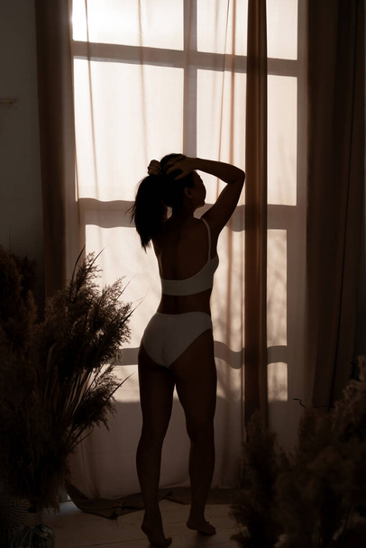 Mujer sexy con cuerpo delgado en ropa interior de pie cerca de la ventana, estirándose cerca de la cama después de despertarse con el amanecer por la mañana, vista trasera - Foto, Imagen