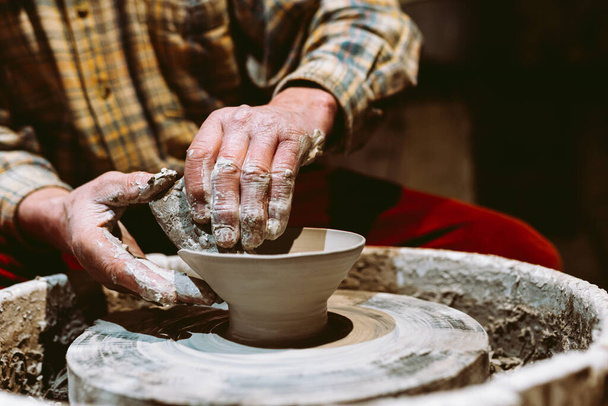 close-up male hands are stained with clay, they are sculpting pottery on potter's wheel. Close-up, soft focus, against blurred background of pottery workshop. Potter's hands at work - Fotografie, Obrázek