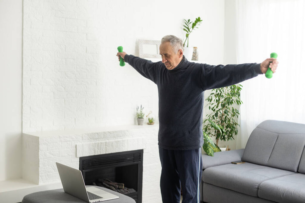 Deporte en edad madura. Feliz hombre de mediana edad haciendo ejercicios de estiramiento delante de la computadora portátil en casa, viendo tutoriales en línea, en el interior de la sala de estar. - Foto, imagen