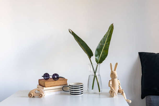 Still life with a wooden hare on hinges in the interior. A vase with two green palm fronds, a white and black striped cup, a stack of books, and a wooden children's toy car. Copy space - Фото, зображення