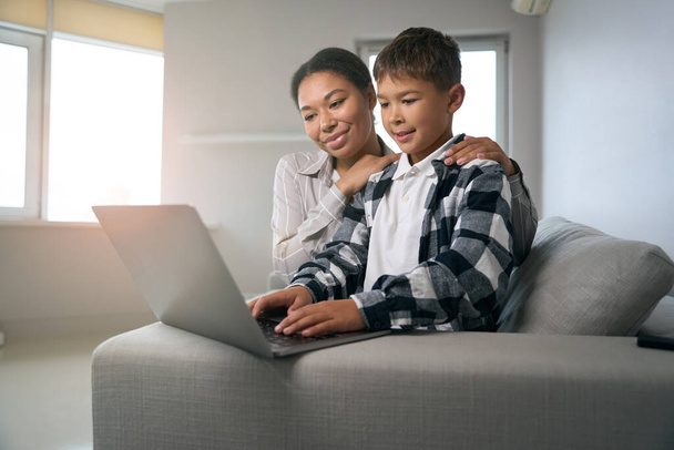 Mom and son are sitting on the sofa with a laptop, a caring woman helps her son in online learning - Photo, Image
