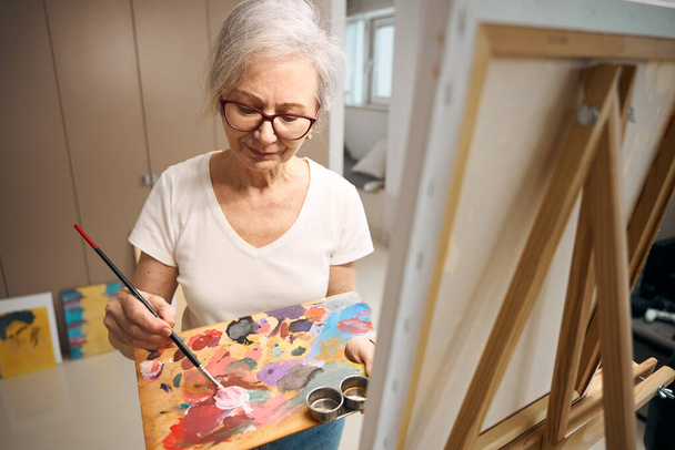 Beautiful elderly lady mixes paints on a palette, a woman paints a picture - Φωτογραφία, εικόνα