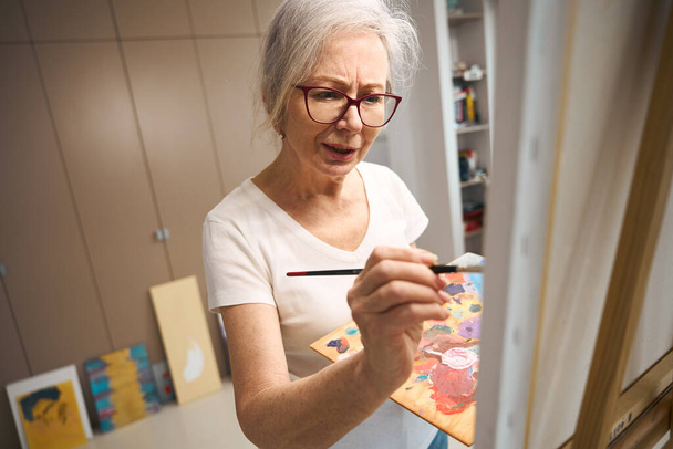 Talented elderly lady paints a picture in a bright room, around her previous works - Φωτογραφία, εικόνα