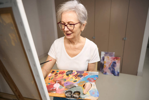 Pensioner holds a palette for drawing in her hands, she stands in front of an easel - 写真・画像