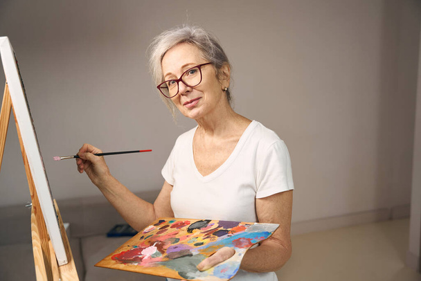 Gray-haired sophisticated lady in glasses stands in front of an easel, she paints a picture - Foto, immagini