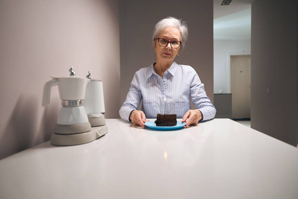 Lonely pensioner sits at a table with piece of cake, on the table there is a set of electric kettles - 写真・画像