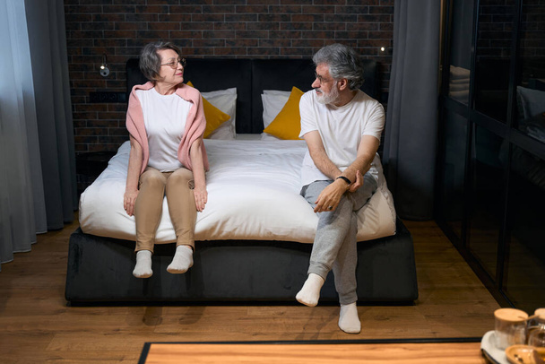 Elderly couple sitting on bed in bedroom, looking at each other, preparing for sleep - 写真・画像