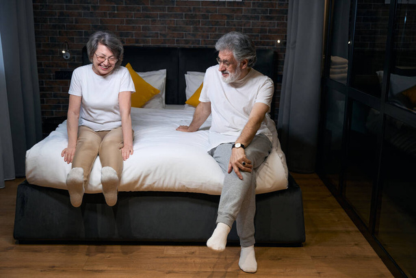 Old happy couple sitting on bed in bedroom, talking with each other, preparing for sleep - Фото, зображення