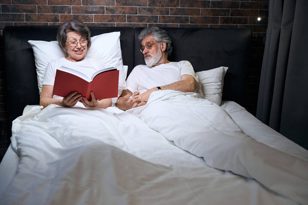 Old lady and man lying on bed in bedroom, wife reading book to her husband before sleep - Valokuva, kuva