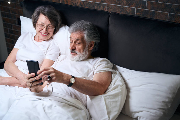 Elderly lady and male lying on bed in bedroom, male holding mobile phone, having video call - Foto, Bild