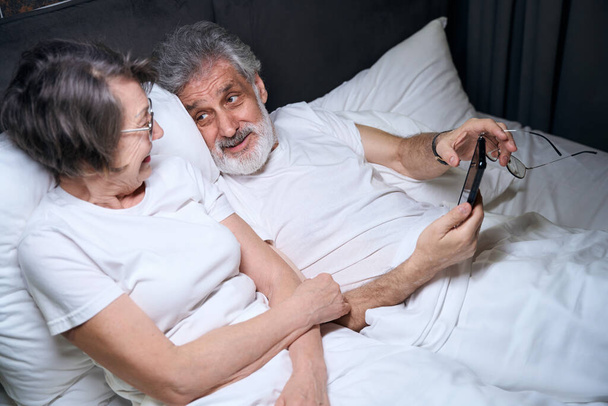 Elderly woman and male lying on bed in bedroom, male holding mobile phone, looking at wife - Foto, afbeelding