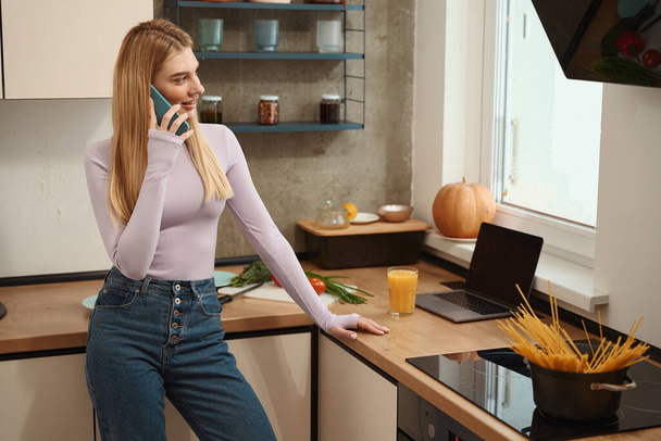 Smiling young woman leaning on kitchen counter and talking on mobile phone - Foto, Imagem