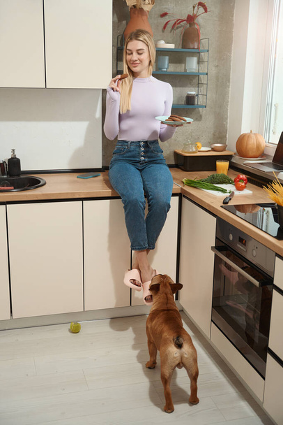 Smiling female seated on kitchen counter looking at oatmeal cookie in her hand - Photo, image