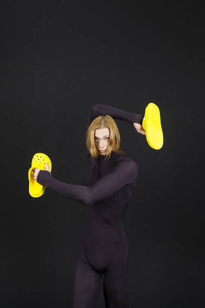 Νεαρός αθλητικός επαγγελματίας χορευτής μπαλέτου σε μαύρο unitard είναι σε τέλεια κατάσταση και ποζάρουν με φωτεινά κίτρινα παντόφλες στα χέρια του πάνω από ένα μαύρο φόντο. - Φωτογραφία, εικόνα