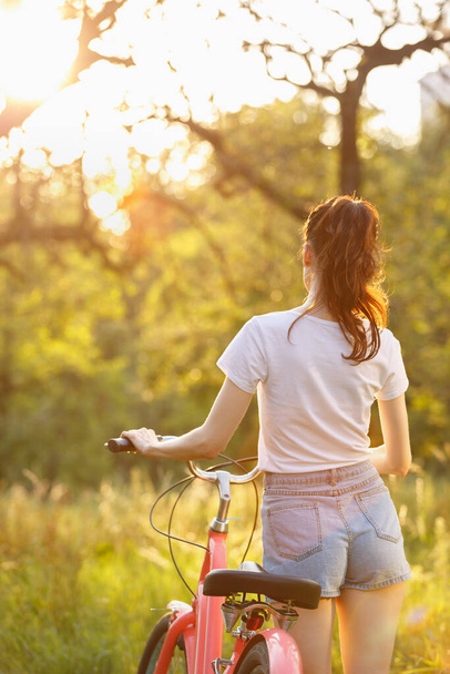 Sexy young woman in shorts walking with a bicycle at sunset. Beautiful white female riding on a bike outdoor in sunny spring day - 写真・画像