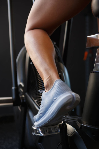 Young woman training on elliptical trainer machine in gym. Close up photo of athlete calf muscle. Athletic female exercising on orbitrek  - 写真・画像