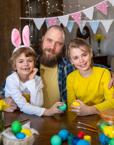 Easter Family traditions. Dad and two caucasian happy children with bunny ears play and decorate eggs with paints for holidays while sitting together at home table having fun together - Foto, Imagen