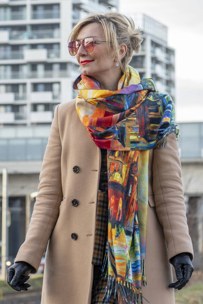 A beautiful and smiling blonde 40-45 years old in a coat and gloves against the background of urban multi-storey buildings. - Fotografie, Obrázek