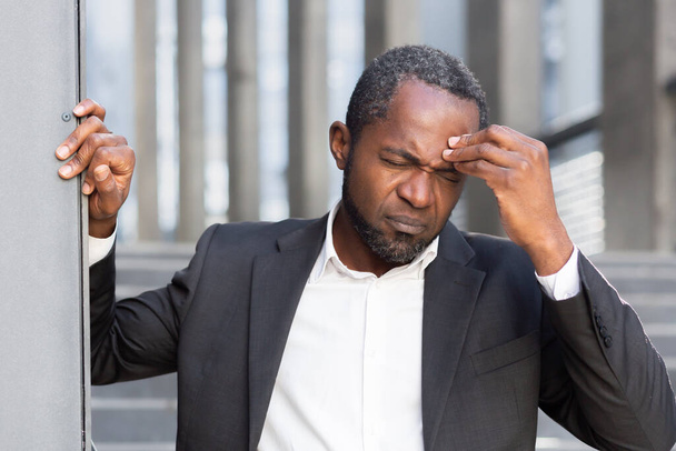 Senior African American man suffering from severe headache, pressure. He is standing outside the office center, leaning against the wall, holding his head, grimacing in pain. - 写真・画像